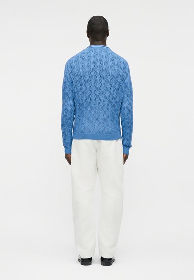 Homme debout, de dos, portant un pull bleu texturé, un pantalon blanc et des chaussures noires, sur un fond blanc uni.