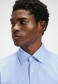 Randig blå skjorta med spetskrage, knäppning och slät tygtextur. Har smala vita ränder och en tajt passform.