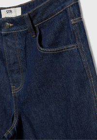 Jean en denim bleu foncé avec poche avant, passants de ceinture, bouton en métal et surpiqûres jaunes visibles.