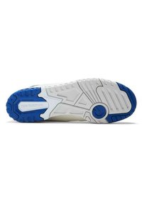 White athletic shoe outsole features a textured surface with blue accents, distinct tread patterns, and a circular emblem near the heel.