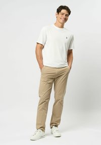 Jaunas vyras šypsosi, rankos kišenėse, dėvi baltą trumpomis rankovėmis marškinius, smėlio spalvos kelnes ir baltus sportbačius, stovėdamas priešais vientisą foną.