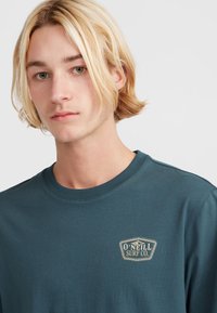 Mėlynas medvilninis marškinėliai su apykakle, turintys išsiuvinėtą O'Neill Surf Co. etiketę ant kairiojo krūtinės, lygus audinys ir laisvas fasonas.