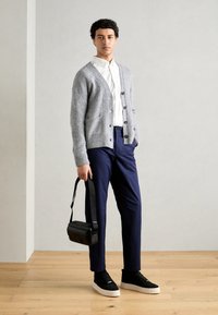 Grå strikket cardigan med knapper, hvit skjorte under, marineblå bukser, svarte sneakers med hvite såler, og en svart crossover-veske.