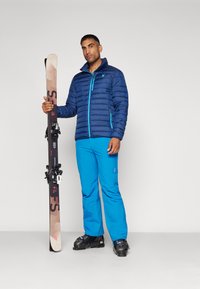 Veste rembourrée navy avec des accents turquoise, pantalon de ski bleu et bottes de ski noires. Tient des skis avec des fixations noires, le dessous beige visible.