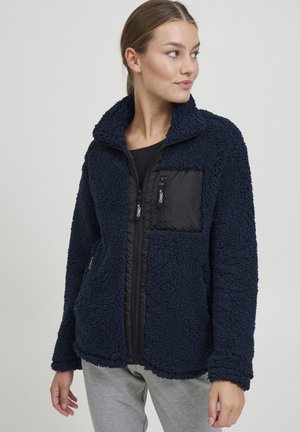 Giacca in pile navy con tasca sul petto trapuntata nera, chiusura frontale completa e tasche laterali. Tessuto morbido e testo con colletto alto.