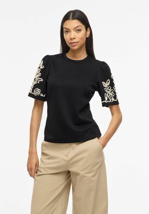 Kvinde med langt mørkt hår iført en sort kortærmet top med beige blomsterbroderi og beige bukser med vide ben, stående med den ene hånd i lommen.