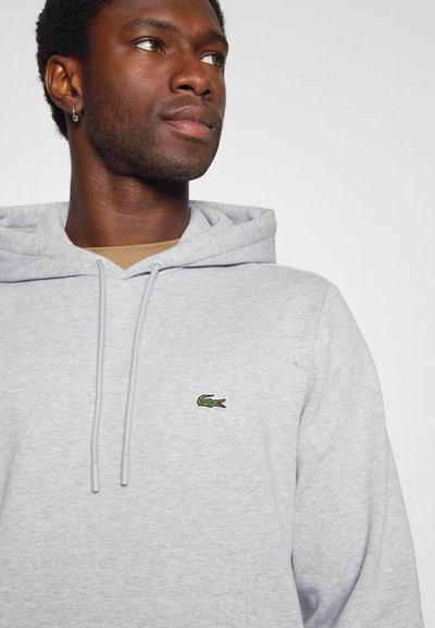 Haut à capuche gris avec une poche kangourou, un capuchon avec cordon de serrage et un petit logo de crocodile vert sur la poitrine. Tissu doux avec une coupe décontractée.