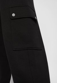 Gros plan d’un pantalon noir avec une poche latérale rectangulaire sécurisée par un rabat et un bouton argenté sur la cuisse.