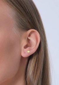 Gul skalformet stud ørestik set på et øre. Glat tekstur med en subtil glans, lille størrelse og detaljeret skaldesign.