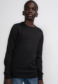 Zwarte crewneck trui, gebreide textuur, lange mouwen, aansluitende boorden en zoom. Gepaard met blauwe spijkerbroek, staand tegen een witte achtergrond.