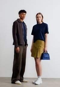 Άνδρας με σκούρο σακάκι, μπλε πουκάμισο και καφέ παντελόνι στέκεται δίπλα σε γυναίκα με μπλε πουκάμισο, κίτρινη καρό φούστα, λευκά αθλητικά παπούτσια, κρατώντας μικρή μπλε τσάντα.