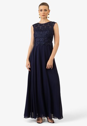 Abito da sera senza maniche di colore blu navy con bustier in pizzo. Gonna fluida con texture a strati. Modella in posa con accessori minimali.