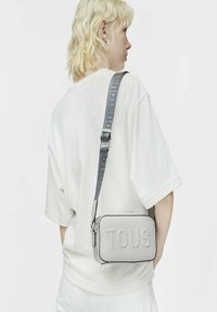 Szara prostokątna torebka z wytłoczonym logo "TOUS", teksturowany materiał oraz szary pasek na ramię z nazwą marki. Noszona w stylu crossbody.