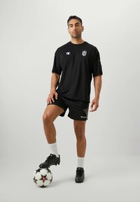 Čierne tričko s logom Champion, čierne šortky, biele ponožky po členky, čierne futbalové topánky a futbalová lopta pri jeho nohách. Jednoduchý športový dizajn.