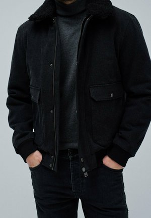 Mann trägt schwarze Reißverschlussjacke mit Lammfellkragen, dunkelgrauen Rollkragenpullover und schwarze Jeans, Hände in den Taschen, vor einfachem Hintergrund.