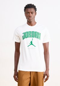 Hvid bomulds t-shirt med stort grønt tekst der står "JORDAN" og et grafisk billede af en springende figur, korte ærmer og en rund halsudskæring.