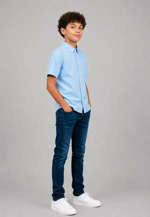 Adolescent aux cheveux bouclés portant une chemise à manches courtes bleu clair, un jean bleu foncé et des baskets blanches, debout les mains dans les poches.