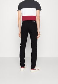 Černé slim-fit džíny v kombinaci s tričkem s barevnými bloky, které má šedé, bílé a burgundské horizontální pruhy. Bílé tenisky s červenými akcenty.