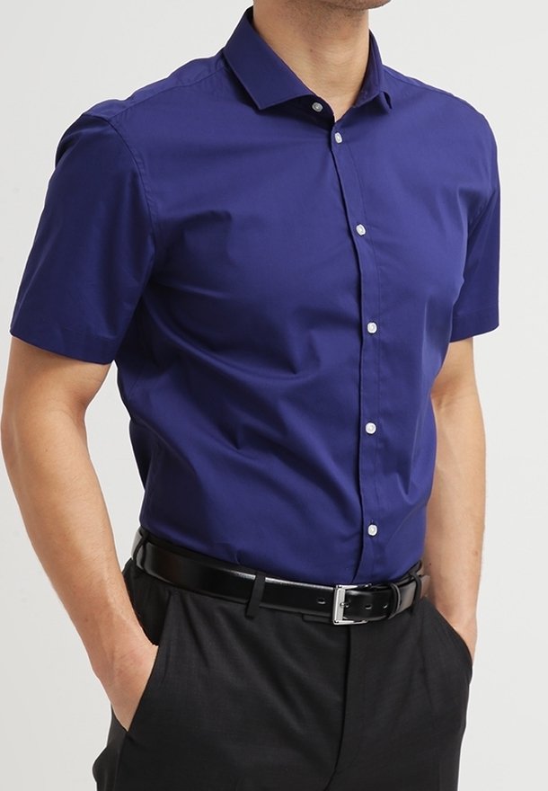 Homme portant une chemise violette à manches courtes boutonnée rentrée dans un pantalon noir avec une ceinture noire, les mains dans les poches.