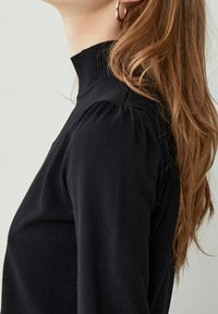 Svart höghalssweater i mjukt material med plissédetalj på axlarna. Har ribbad krage och slät textur.