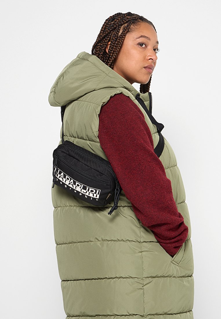 Osoba z warkoczami nosi oliwkową kurtkę puchową, bordową bluzkę z długim rękawem i czarną torbę na ramię Napapijri, patrząc na bok.