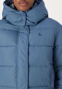 Giacca trapuntata azzurro chiaro con colletto alto, caratterizzata da un motivo cucito e un logo sul lato sinistro del petto. Tessuto morbido e liscio.