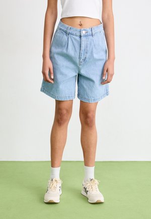 Persoon draagt lichtblauwe denim shorts, wit cropped top, witte sokken en beige sneakers, staat op een groene vloer tegen een witte muur.