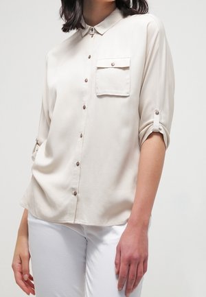 Vrouw met een beige overhemd met opgerolde mouwen en een borstzak, gecombineerd met een witte broek, tegen een effen achtergrond.