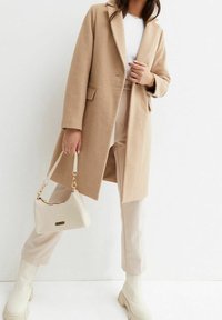 Manteau en laine beige avec un col cranté, associé à un pantalon beige clair et des bottes blanches mi-mollet. Tient un sac à main matelassé blanc avec des accents dorés.