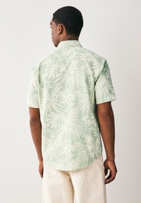 Camicia a maniche corte verde chiaro con motivo a foglia di palma bianca. Presenta un colletto abbottonato e un orlo curvo. Tessuto testurizzato.
