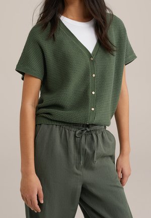 Une femme porte un cardigan en tricot olive à manches courtes boutonné par-dessus une chemise blanche, avec un pantalon vert olive à cordon de serrage, sur un fond beige.