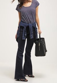 Mujer con una blusa de manga corta estampada, jeans oscuros acampanados, sandalias, con una chaqueta de mezclilla atada a la cintura, sosteniendo un bolso negro.