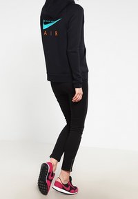 Svart Nike-hoodie med turkos swoosh och orange "AIR"-text på baksidan. Kombinerad med svarta slim fit-jeans och rosa Nike-sneakers.