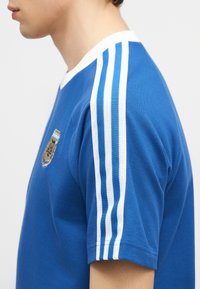 Πλάγια όψη προσώπου που φοράει μπλε αθλητική φανέλα με λευκές ρίγες στον ώμο και ένα μικρό έμβλημα στο στήθος.