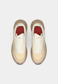 Une paire de chaussures de course beiges et brunes avec des lacets blancs et des semelles intérieures rouges, vue de dessus sur un fond blanc.