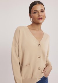 Beżowy sweter z dzianiny z dekoltem w kształcie litery V, sześcioma guzikami i dwiema kieszeniami z przodu. Posiada prążkowane mankiety i dół, dodające faktury.