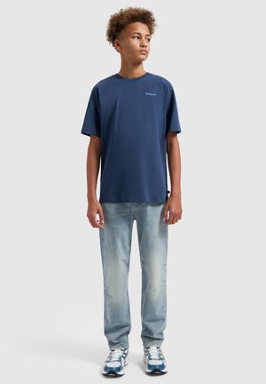 Tienerjongen staand, draagt een marineblauw t-shirt, lichtblauwe spijkerbroek en wit-blauwe sneakers, kijkt iets naar de zijkant.