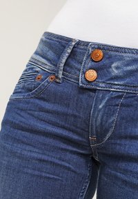 Närbild av en person som bär blå denimjeans med kopparknappar och nitar, tillsammans med en vit topp.