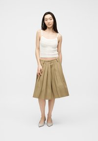 Νεαρή γυναίκα με λευκό αμάνικο τοπ από πλεκτό, μπεζ πλισέ midi φούστα και μπεζ ψηλοτάκουνες γόβες με μυτερή μύτη, που στέκεται μπροστά σε μονόχρωμο φόντο.