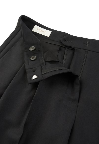Czarne spodnie z plisowanym przodem, z zapięciem na zamek błyskawiczny, dwoma czarnymi guzikami i metką z napisem „SLOWEAR”. Gładka struktura materiału, dopasowany fason.