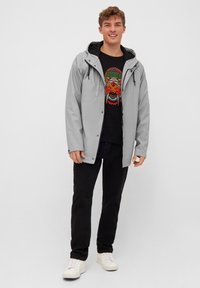 Light gray waterproof jacket with a hood, featuring a black lining. Paired with a black shirt displaying a colorful graphic design and dark pants.
