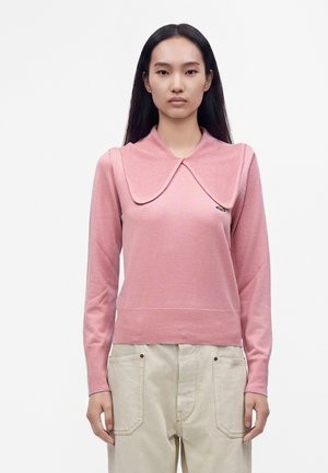 Femme aux longs cheveux noirs portant un haut rose à manches longues avec un col oversize et un pantalon beige clair, regardant vers l'avant sur un fond uni.