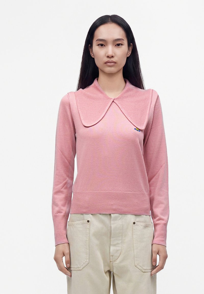 Femme aux longs cheveux noirs portant un haut rose à manches longues avec un col oversize et un pantalon beige clair, regardant vers l'avant sur un fond uni.