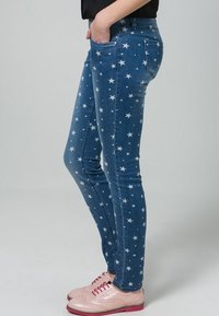 Jean en denim bleu ajusté avec un motif d'étoiles blanches, comportant cinq poches et une taille mi-haute. Assorti à des chaussures à lacets rose pâle.