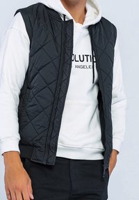 Homme portant un sweat à capuche blanc avec le texte "Los Angeles" partiellement visible, un gilet sans manches noir matelassé ouvert, et un pantalon noir devant un fond uni.