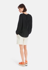 Schwarze Bluse mit langen Ärmeln, lässigem Schnitt und gefaltetem Rücken, kombiniert mit cremefarbenen Shorts und orangenen Sandalen mit geflochtenen Details.