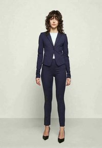Fato azul-marinho sob medida com um blazer ajustado e calças slim taper, combinado com uma blusa branca e sapatos de salto alto pretos. Tecido suave, design minimalista.