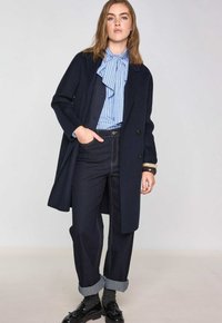 Cappotto di lana blu navy con una vestibilità su misura, indossato sopra una blusa a righe blu chiaro. Un paio di jeans scuri con risvolti e scarpe nere.