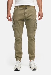 Uomo che indossa pantaloni cargo verde oliva con polsini elasticizzati, cintura abbinata, sneakers bianche e t-shirt bianca su sfondo semplice.