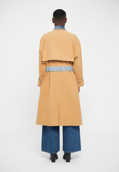 Manteau trench en coton beige avec des accents en denim, présentant un dos en couches, des manches larges et une ceinture à la taille. Porté avec un jean bleu à jambe large.